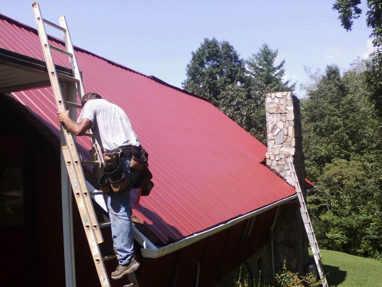 Roofer on a ladder beside a red metal roof and stone chimney — Soffit & Fascia Repair by Roofing Brothers in Lake Hopatcong